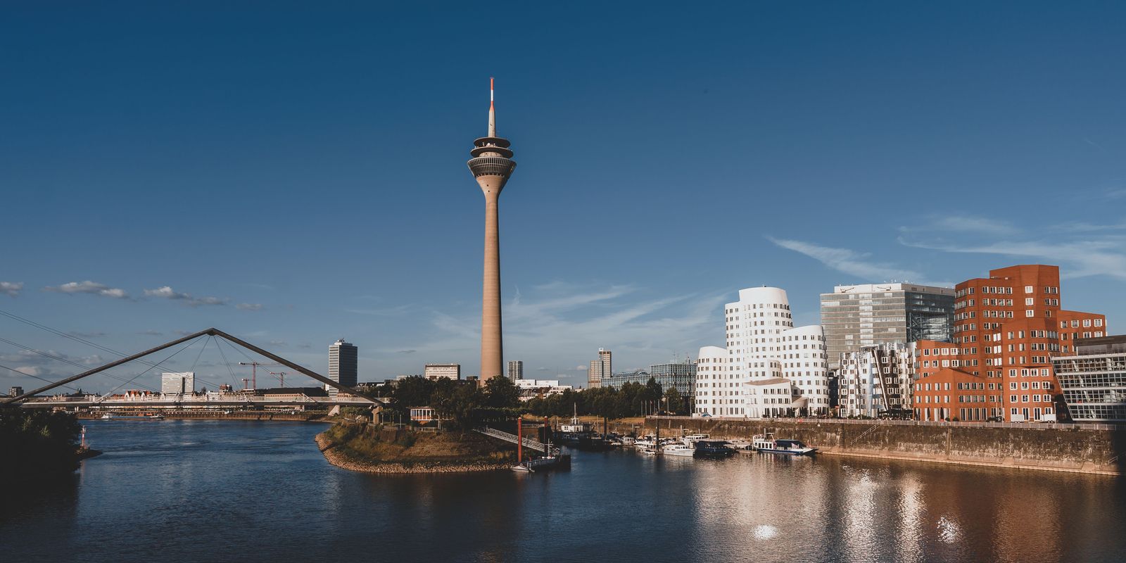 Düsseldorfer Medienhafen mit Rheinturm, den geschwungenen Gehry-Bauten und dem Hafenbecken unter blauem Himmel